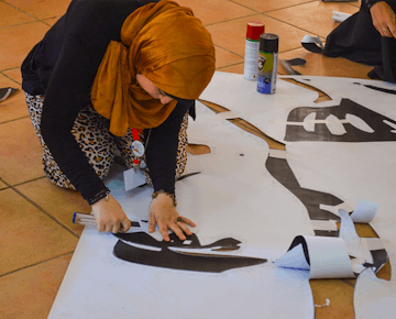 A woman in a mustard headscarf carefully cuts a large stencil design on the floor, surrounded by art supplies and spray paint.