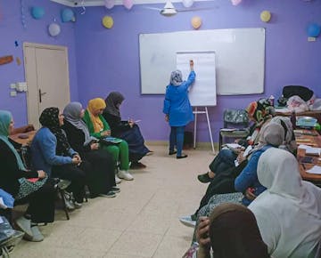 A group of women sit in a purple classroom while an instructor writes on a flip chart, facilitating a workshop in a supportive learning environment.