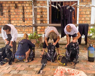 A group of four marine conservation volunteers are bent over checking and assembling scuba diving equipment in an outdoor courtyard with hanging wetsuits in the background.