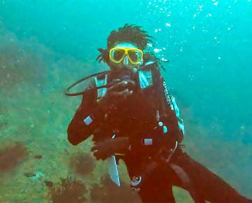 A scuba diver in full gear is underwater, facing the camera and holding their breathing regulator. The diver wears a black wetsuit and yellow diving mask, surrounded by clear blue water and the rocky, plant-covered seabed typical of Mozambique's marine environment.