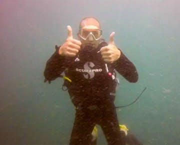 A scuba diver underwater, wearing a black wetsuit and dive mask, faces the camera and makes the double Thumbs Up hand signal.