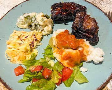 A dinner plate includes grilled meat, pap (maize meal), tomato sauce, cooked greens, coleslaw, and fresh salad, served to IVHQ volunteers in Mozambique.