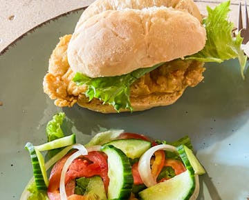 A breaded chicken burger with lettuce on a soft bun is paired with a simple salad of tomato, cucumber, and onion, served as a volunteer lunch in Mozambique.
