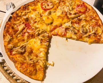 A freshly made pizza with toppings like chicken, tomato, and cheese is partially sliced and served on a white plate. An example of a meal purchased in Mozambique.