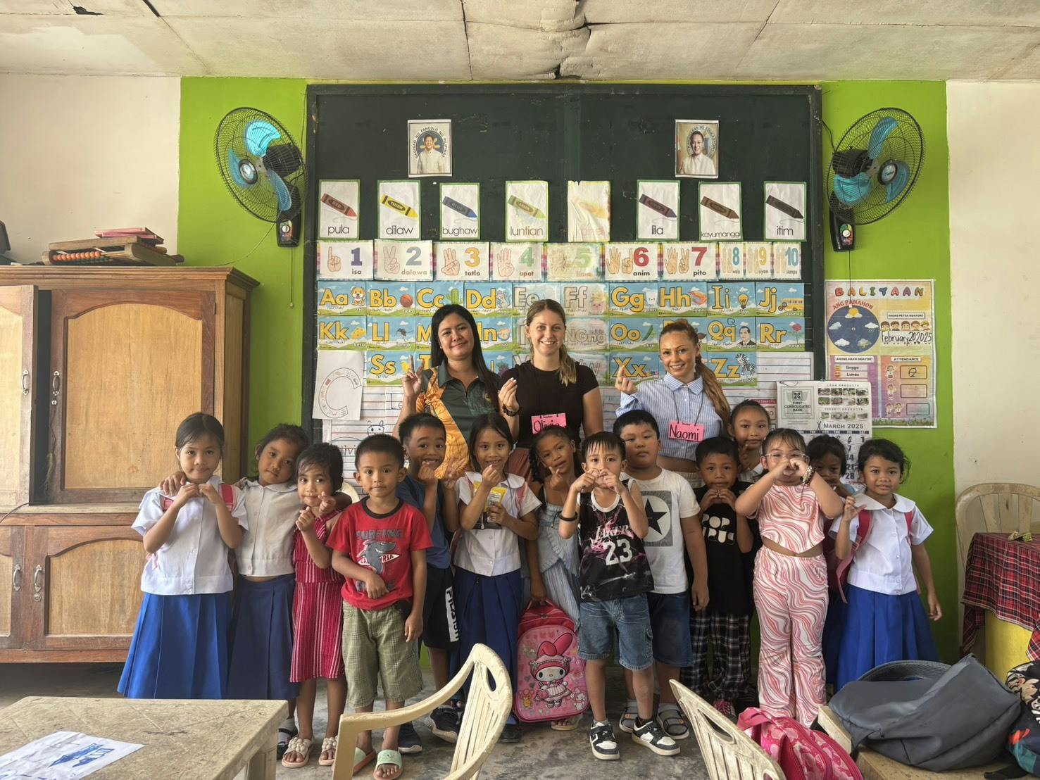 Two young female volunteer teachers stand at the front of the class, smiling with students.