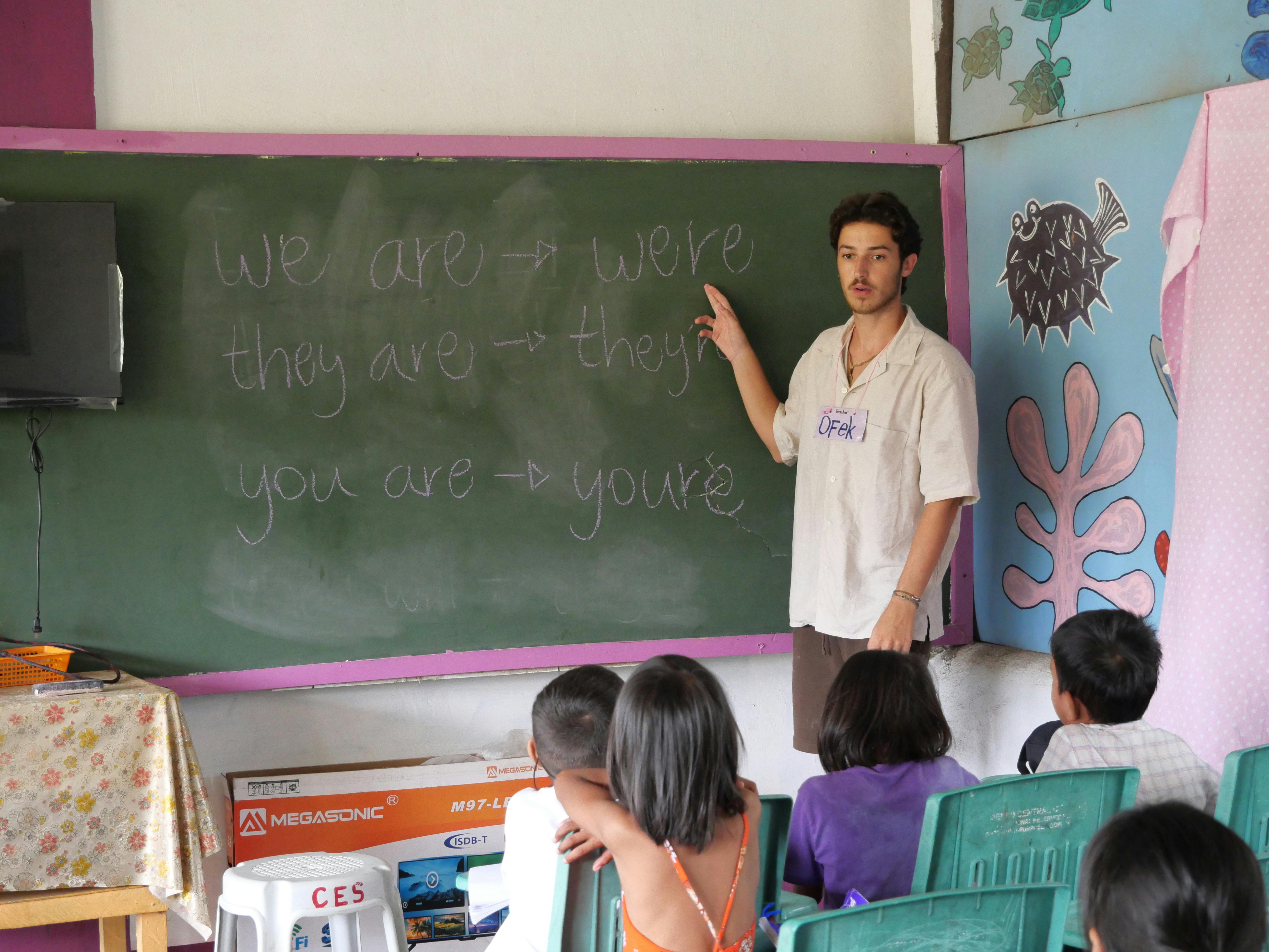 An international teaching volunteer stands at the front of a classroom, teaching from the chalkboard as rows of young students listen and participate.