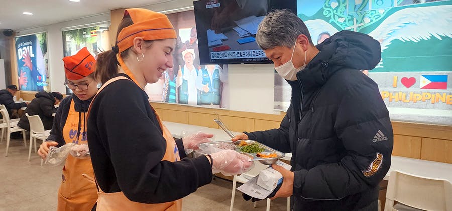 A volunteer passing a plate of set meal to people at the IVHQ Soup Kitchen project in Seoul, South Korea