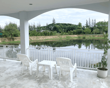 A covered balcony with two white chairs and a table, overlooking a peaceful pond and green landscape.
