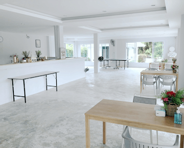 A large, airy communal space with tables, chairs, plants, and a countertop, all in a minimalist white decor.