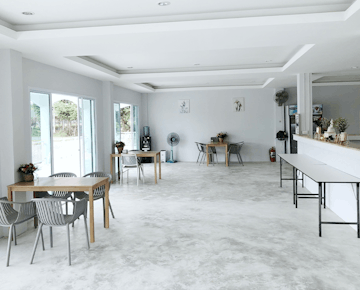 A spacious, modern common area with white walls, light wooden tables, gray chairs, potted plants, and large windows letting in natural light.