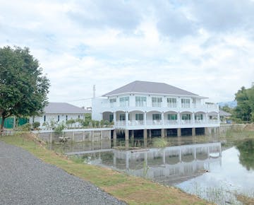 A bright, modern accommodation building on stilts, reflected in a pond, surrounded by gardens and trees.