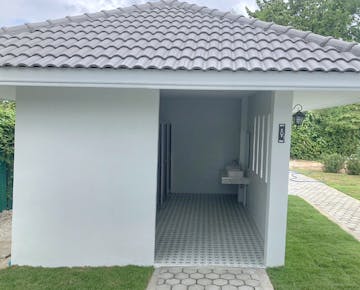 A small, white-tiled building with a sloped roof, open to reveal a clean shared bathroom area with sinks and private stalls.