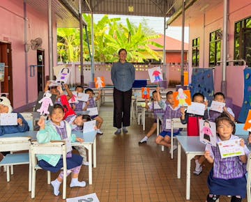 A group of young children in uniform smile and hold up their colorful drawings in a bright classroom, with a volunteer standing at the center.