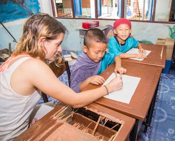 A volunteer sits at a wooden desk, helping two boys in traditional clothing as they write on paper, in a classroom decorated with murals.