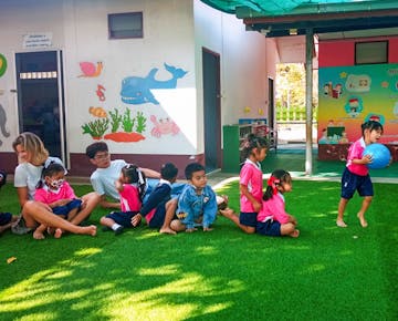 Children and volunteers sit in a line on artificial grass outdoors while a child in pink uniform runs with a blue ball; the background features playful wall art.