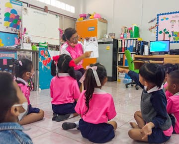 A teacher in a pink uniform shows a drawing to a group of children sitting on the floor, who eagerly raise their hands in a colorful classroom.