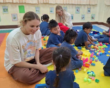 A volunteer sits cross-legged on a mat, smiling and interacting with a child while others play with blocks around them in a classroom.