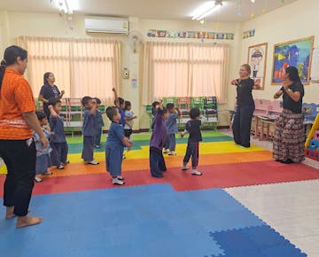 Children and teachers, including a volunteer, stand on bright foam mats and participate in a dance or movement activity in a cheerful classroom.