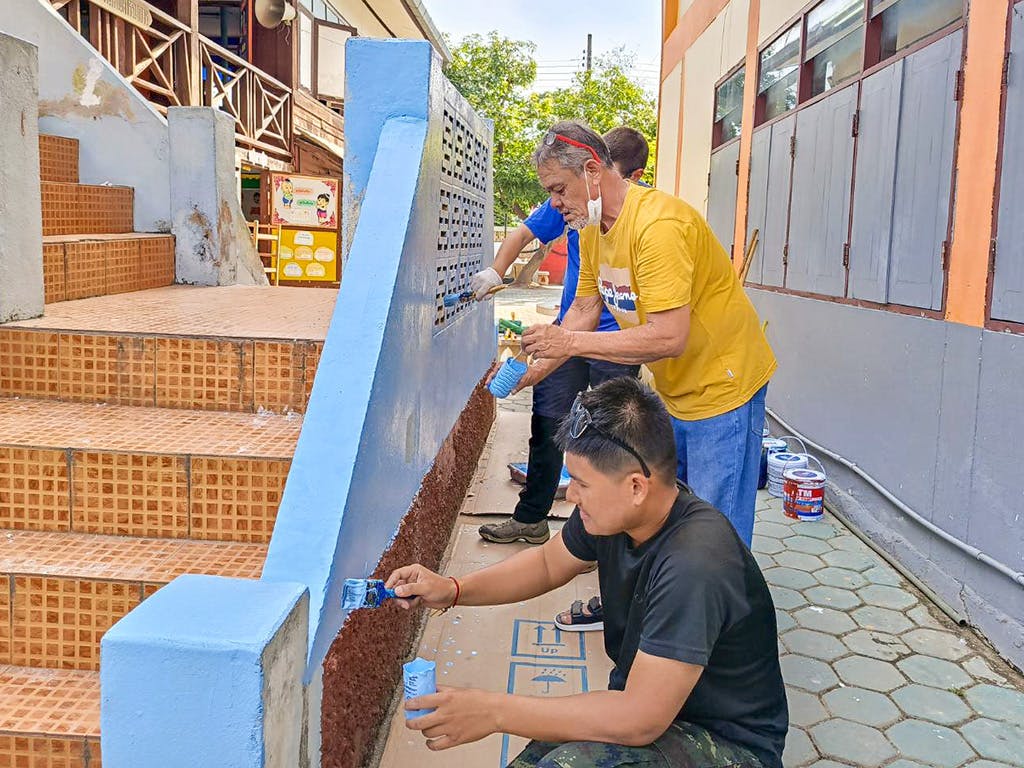 A group of volunteers paint the exterior stairs and wall of a school a bright blue color, working together with brushes and rollers.