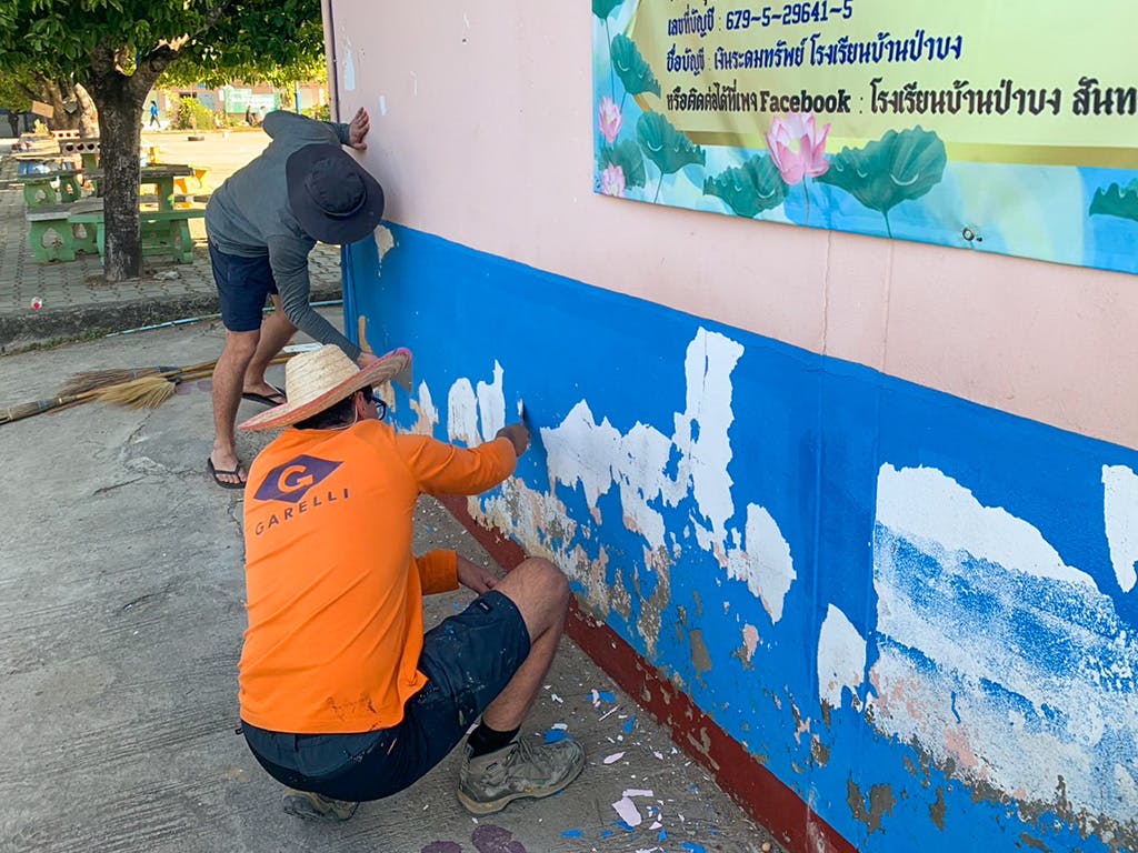 Two volunteers scraping and preparing an exterior wall for a fresh coat of paint, with blue paint peeling off the lower section.