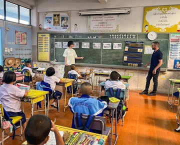 Two international teaching volunteers stand at the front of a classroom, teaching from the chalkboard as rows of young students in uniform listen and participate.