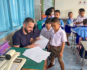 A male volunteer teacher seated at a desk hands out papers to a group of smiling students in white uniforms gathered around him.