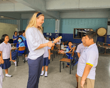 A young female volunteer teacher stands at the front of the class, laughing and holding markers while interacting with smiling students.