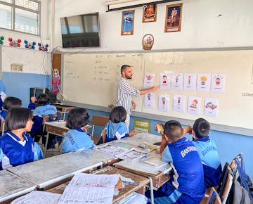 A volunteer teacher points to illustrated cards on the whiteboard, explaining different emotions to a classroom of attentive students.