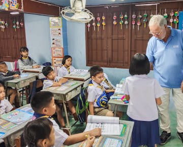 An older male volunteer teacher in a light blue shirt helps a young student at her desk while other primary school children watch in the classroom.