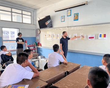 A volunteer teacher points to country flags on the board while a classroom of students in white uniforms looks on.