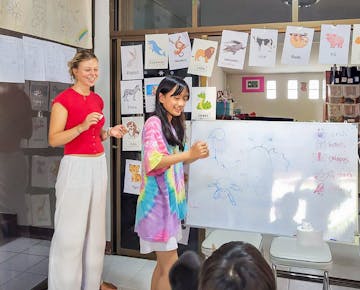 A volunteer and a student stand together at a whiteboard, teaching about animals with colorful illustrated cards on display behind them.
