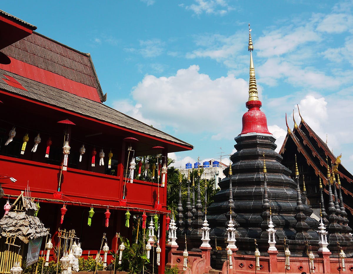 A traditional Thai temple in Chiang Mai with a red building and black-and-red stupa, topped with a golden spire, stands under a bright blue sky with scattered clouds. Decorative lanterns hang along the temple’s exterior.