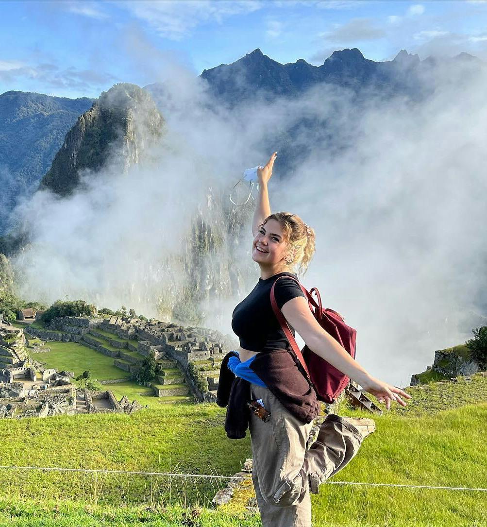 A smiling young female volunteer with a red backpack poses energetically at Machu Picchu, Peru, with mist-covered mountains in the background. She raises one arm and balances on one leg, The ancient Incan ruins below are partially visible through the clouds, adding to the breathtaking scenery.
