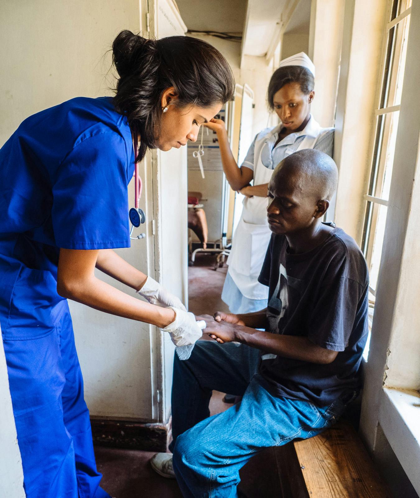 A healthcare volunteer in blue scrubs and gloves providing medical assistance to a seated patient in a clinical setting. The patient, wearing a black t-shirt and jeans, is receiving treatment on his hand. A nurse in a white uniform stands in the background observing.