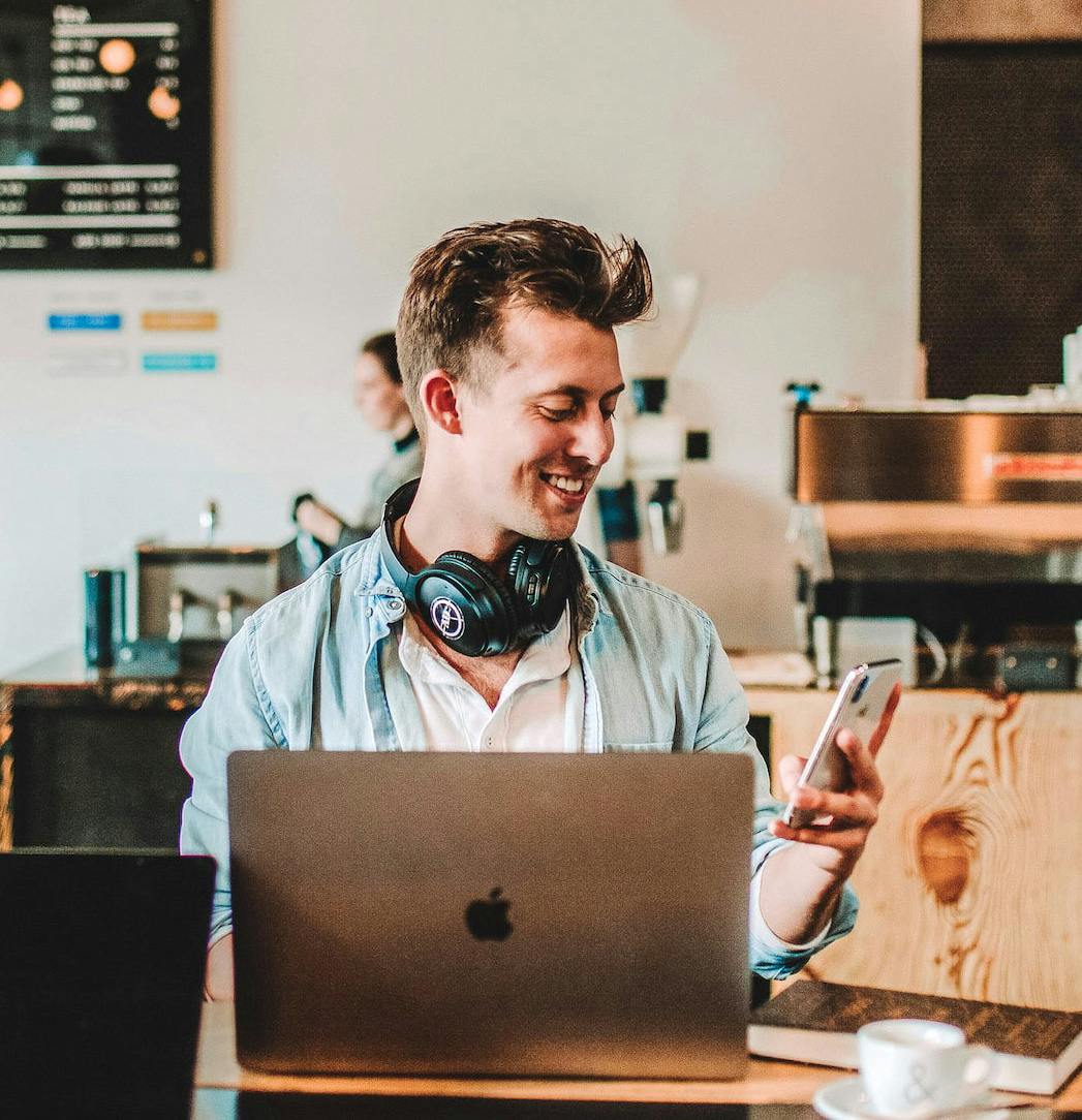 Young man sitting at a café, smiling while looking at his smartphone. He has a laptop open in front of him and a pair of headphones around his neck. The background shows a coffee shop setting.
