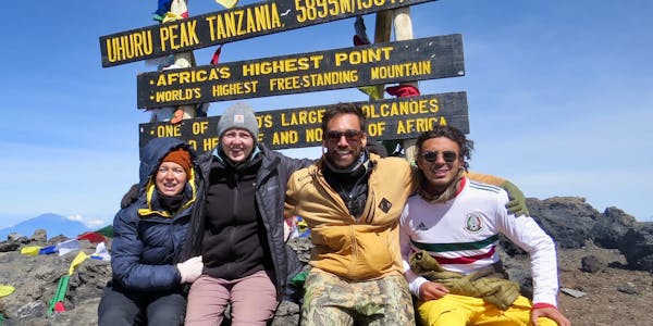 A group of four climbers who are international volunteers sit together, smiling, in front of the Mount Kilimanjaro summit sign at Uhuru Peak, Tanzania, which marks 5,895 meters (19,341 feet) above sea level.