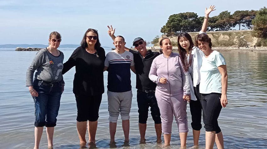 A diverse group of cheerful volunteers and community members from the special needs volunteer project in Croatia stand closely together with feet in shallow water, smiling, waving, and enjoying the sunshine.