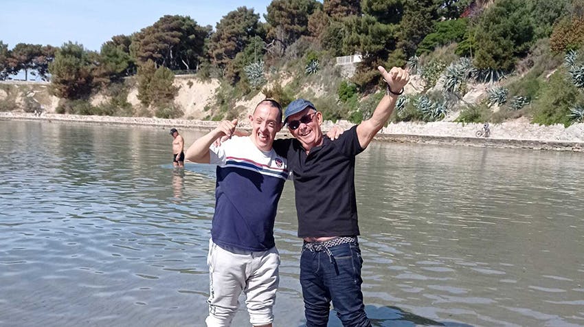 A smiling older volunteer and a joyful young man from the special needs project in Croatia stand side-by-side in shallow, calm water at a beach, both enthusiastically giving thumbs-up gestures.