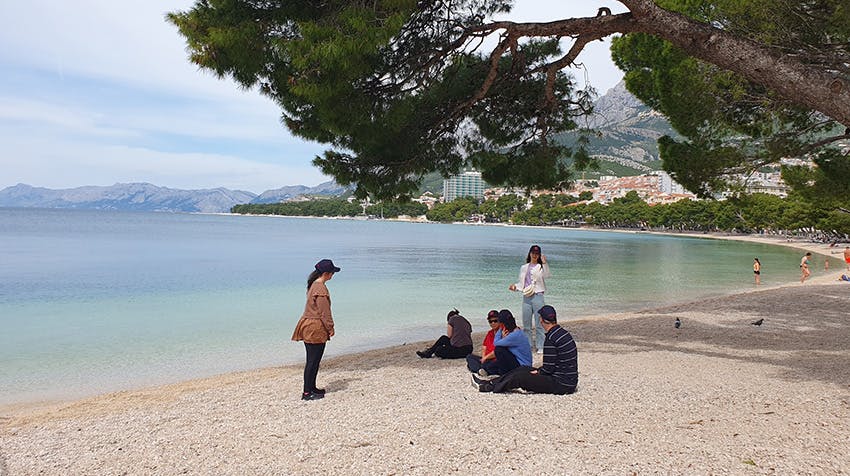Volunteers and community members sitting and standing together on the pebbled shore, shaded by a large tree.