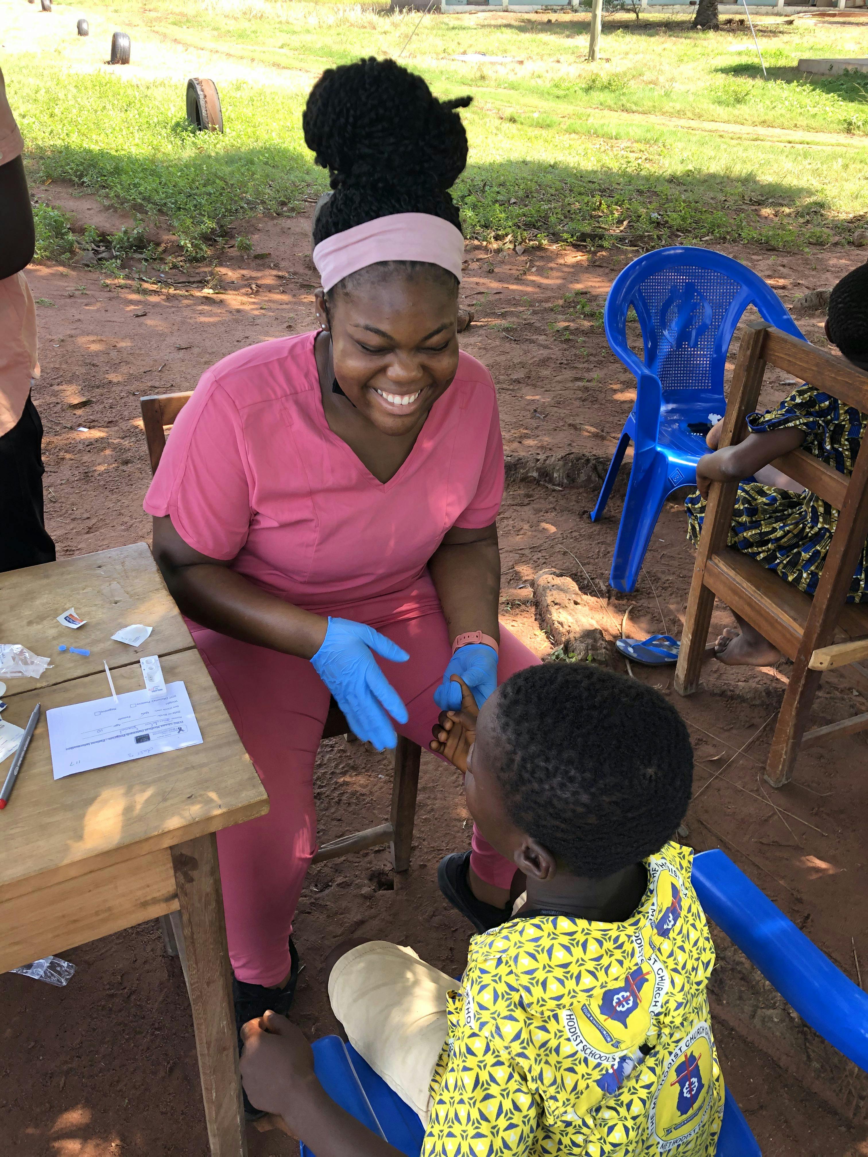 A smiling healthcare volunteer wearing pink scrubs and blue gloves provides medical assistance to a young child sitting in a blue plastic chair.