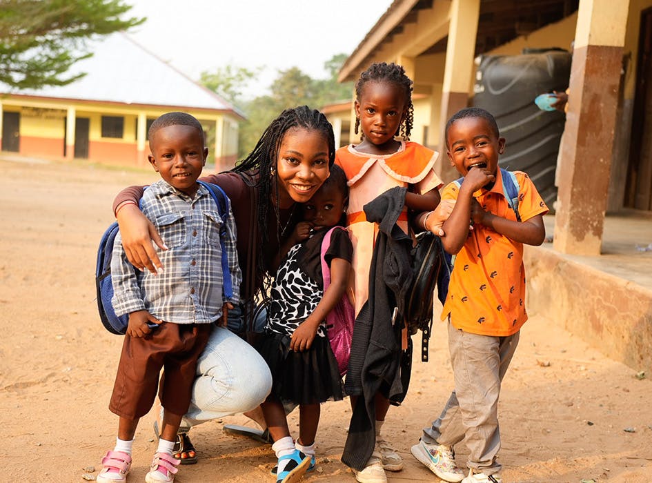 A female mission trip volunteer poses with four children outside a school in a warm, sunny environment. The children, wearing colorful outfits and carrying backpacks, radiate joy, capturing the spirit of community and the positive impact of missionary trip work in education.