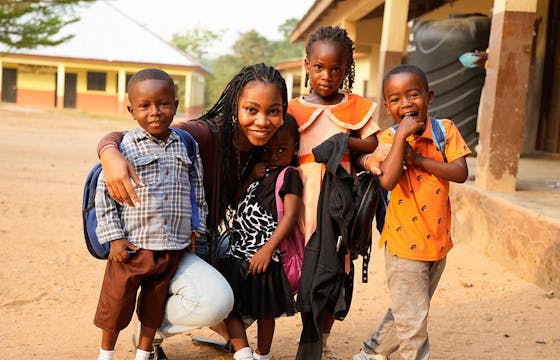 A female mission trip volunteer poses with four children outside a school in a warm, sunny environment. The children, wearing colorful outfits and carrying backpacks, radiate joy, capturing the spirit of community and the positive impact of missionary trip work in education.