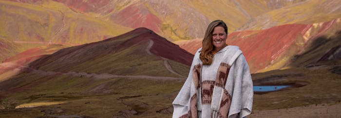 A smiling volunteer traveller wearing a traditional Andean poncho stands in front of the breathtaking Rainbow Mountain in Peru. The landscape features vibrant red, yellow, and green hues across the mountains, with a small lake visible in the background.