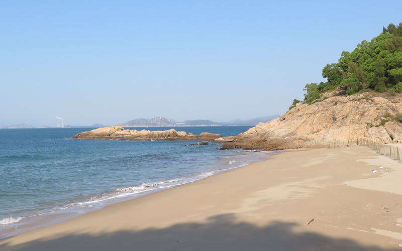 A quiet sandy beach with gentle waves, rocky outcrops, and a hillside covered in green trees under a clear blue sky in Huidong, China.