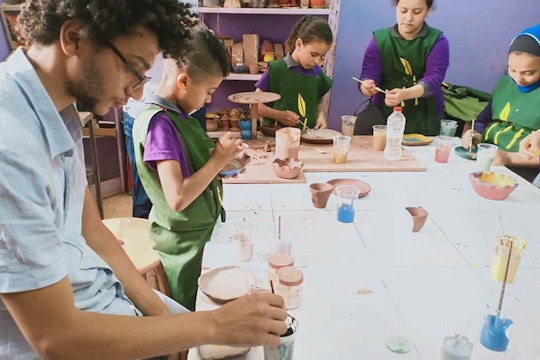 A childcare volunteer in Cairo, Egypt through the IVHQ childcare project and a group of children sit around a table carefully painting clay pottery pieces in a classroom filled with colorful materials and ceramic artworks.