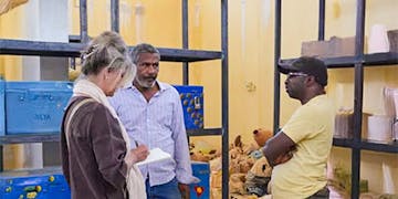 A social media & marketing volunteer in Cairo, Egypt takes notes while speaking with two local men inside a supply storage room, gathering stories and visuals to support marketing and community outreach efforts.