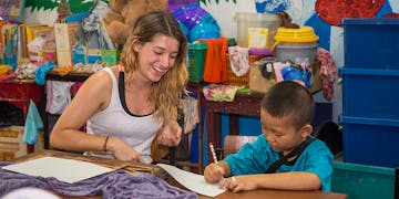 A young female international volunteer smiles warmly as she sits beside a young boy in a preschool in Chiang Mai, Thailand. The volunteer encourages the boy, who is focused on writing with a pencil on a sheet of paper.
