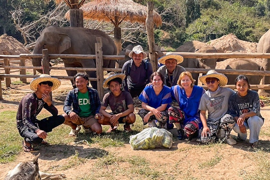 A group of international volunteers and local staff kneel and sit together, smiling for a photo in front of a wooden fence, with elephants and forested hills in the background at an elephant sanctuary in Chiang Mai, Thailand.